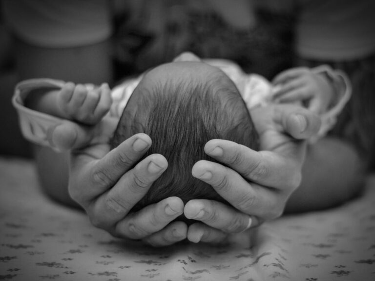 Cabecinha de recém-nascido acolhida entre as mãos de um adulto, foto em preto e branco, sem mostrar o rosto